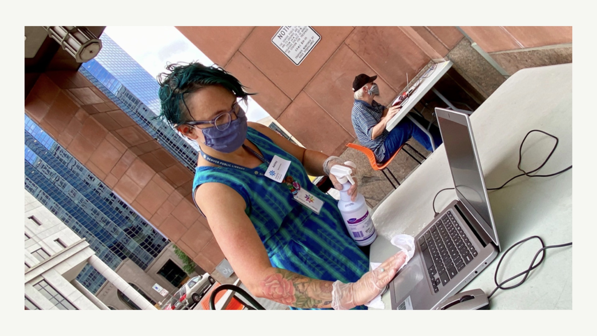 [photograph of Denver public librarian masked and gloved wiping down a public laptop in an outdoor atrium area]