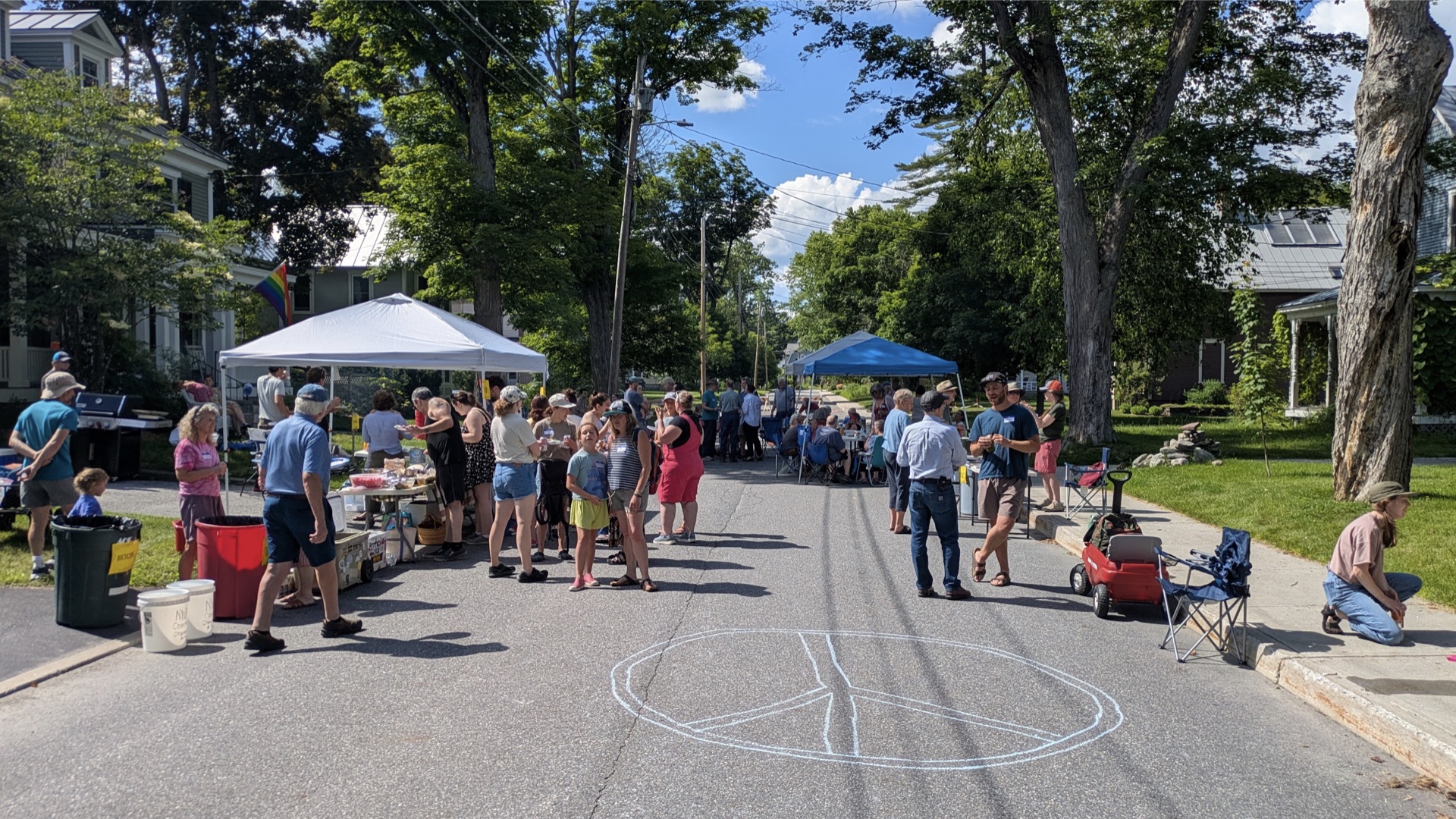 snapshot of a block party with tents set up, a peace sign drawn on the road and many people interacting and talking with one another