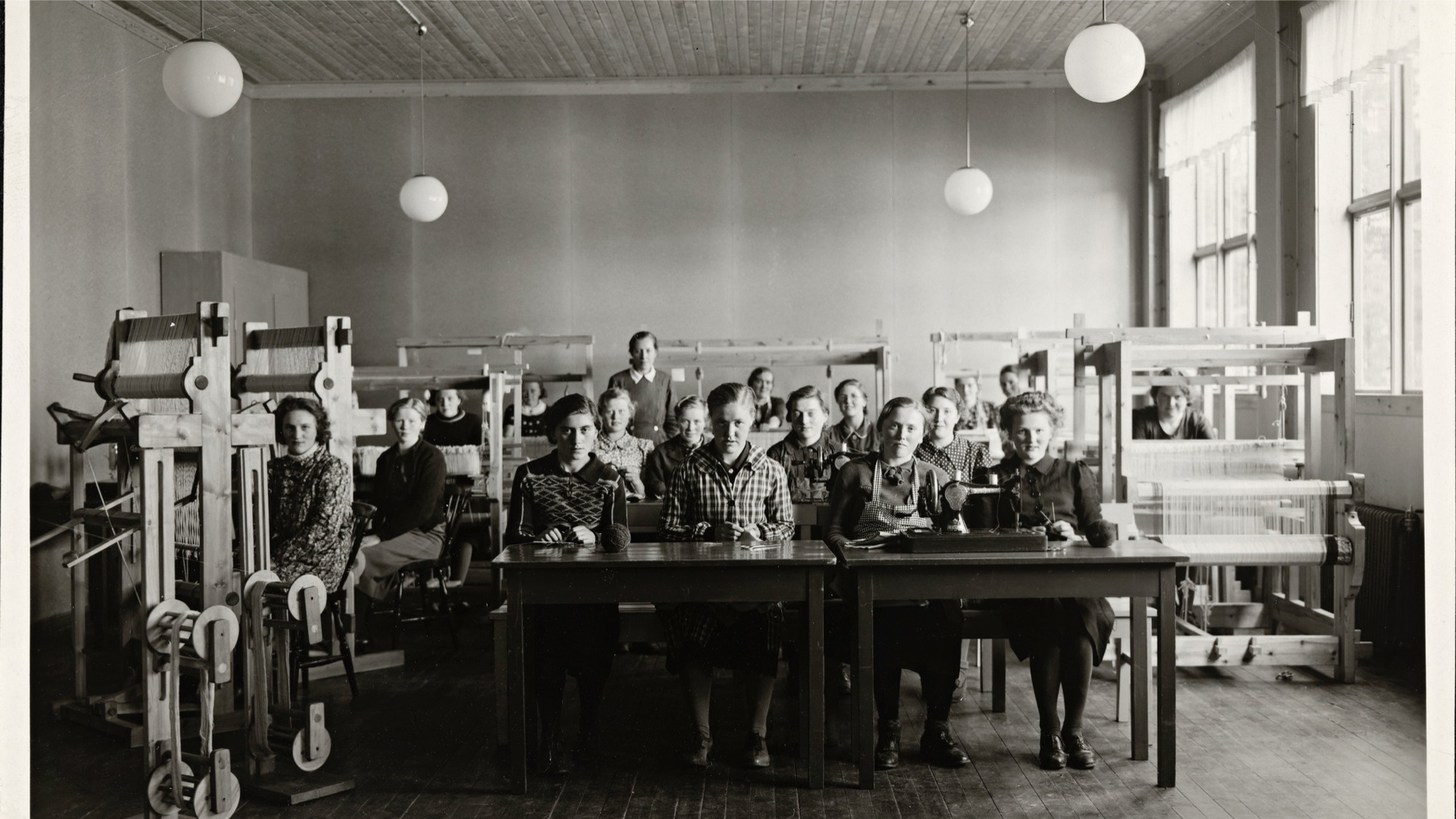 Historical image of a bunch of young women in a room filled with looms.