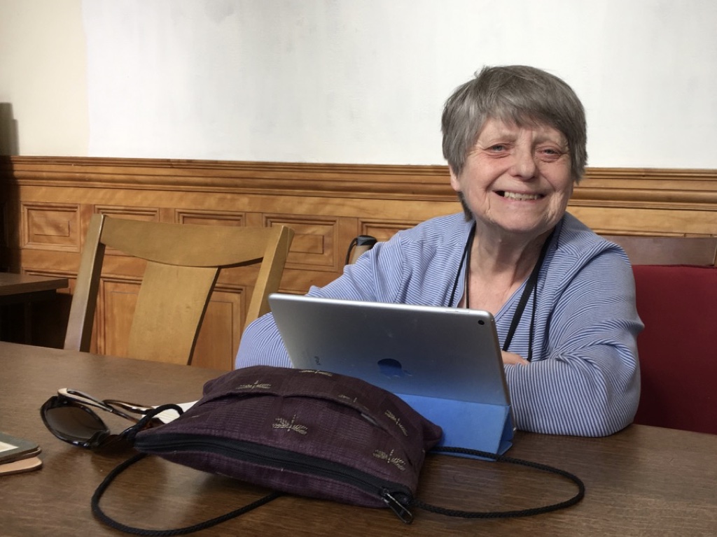Older woman grinning into screen sitting behind her ipad in a wood paneled room