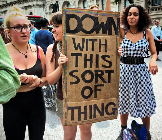 scene of a protest with a woman holding a sign that says "Down with this sort of thing"