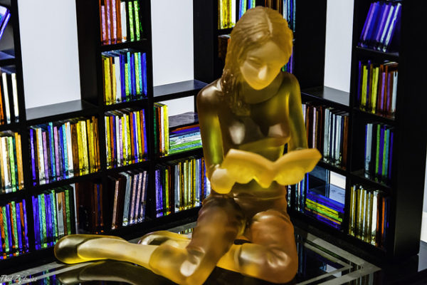 Image of a glass statue of a woman who appears to be reading a book in front of a bunch of illuminated glass books