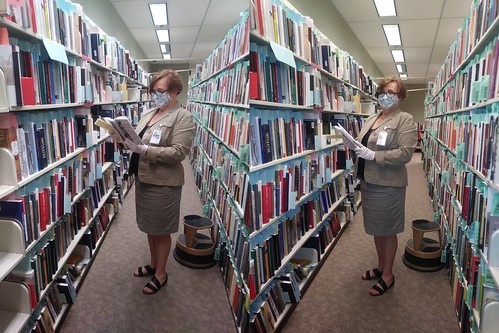 two shots of a woman reading books in a library stacks aisle with her mask on, In oe of the shots she is looking at the book, in another she is looking at the camera