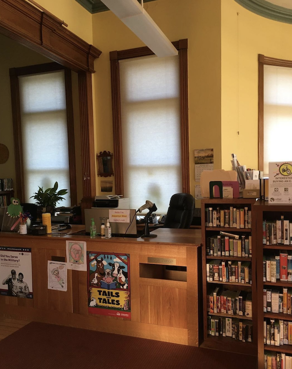 a dim afternoon-into-evening shot of an empty library circ desk with a book drop slot and a small shelf next to it with books on CD