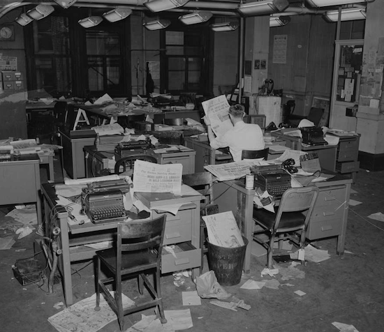 black and white photo of Man sitting in a disarrayed newsroom, with newspapers scattered about