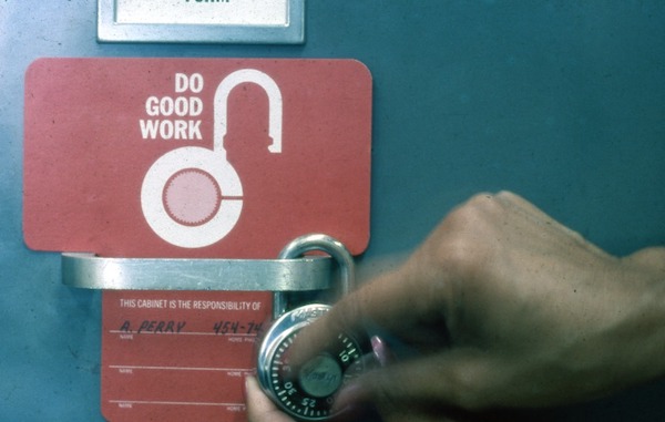 Photograph of a person's hand adjusting a padlock. It's attached to a handle with a sign nearby that says "DO GOOD WORK"