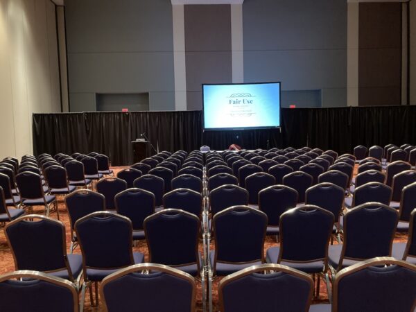a small screen showing a slide that says "Fair Use" inside a large empty ballroom-size room at a convention center