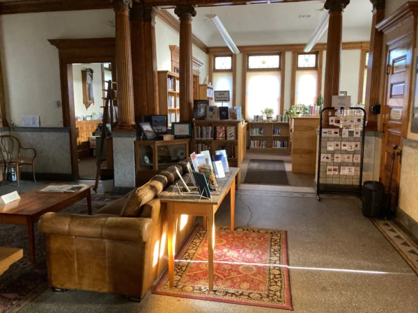 The interior of my public library which has a nice carpet, a leather couch. some stained glass windows, and some bookshelves and wooden columns which are all sort of glowing in the late afternoon sun.